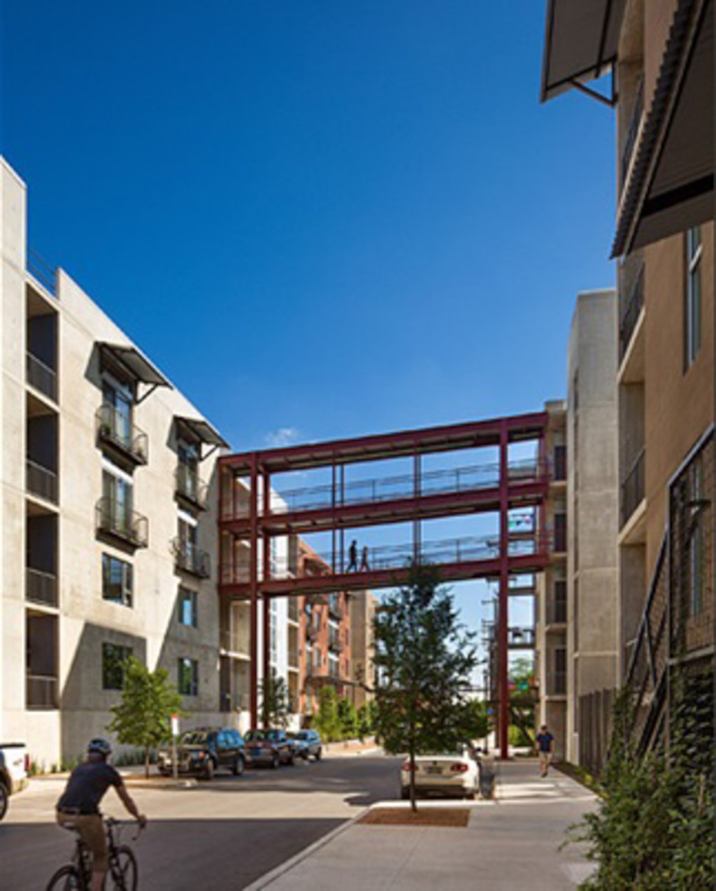 A man rides his bicycle down a sidewalk in front of a building with a large red structure in the background.