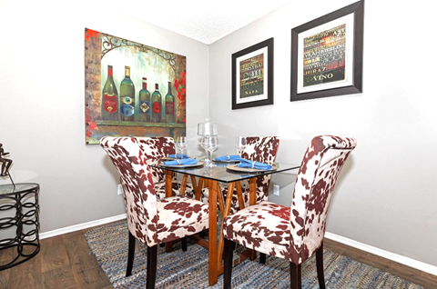 A dining room with a glass table and chairs with a wine bottle painting on the wall.