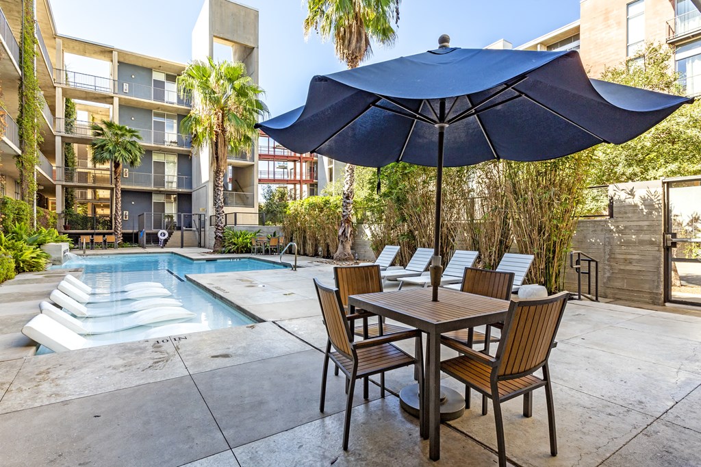 A table with chairs and an umbrella is in the foreground of a pool area.