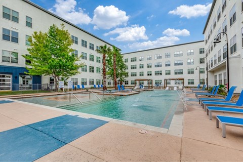 A large swimming pool in front of a building with blue lounge chairs on the side.