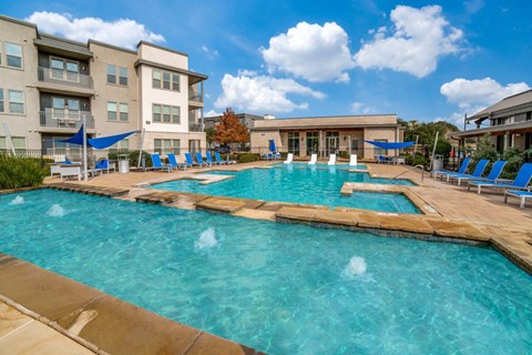 a swimming pool with blue lounge chairs and umbrellas in front of an apartment building at The Landings at Brooks City-Base, Texas