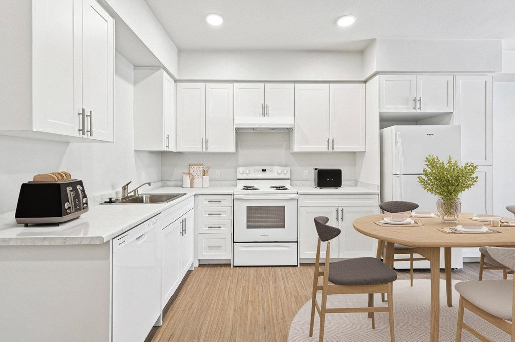 A modern kitchen with white cabinets and appliances, a wooden table with chairs, and a plant on the table.