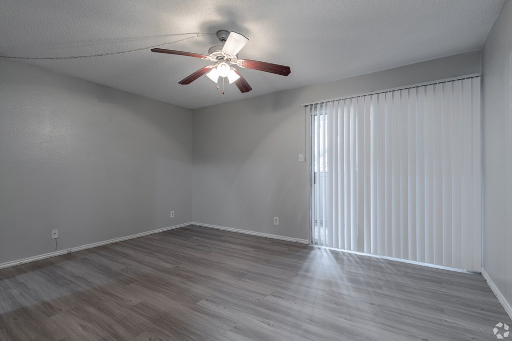 A room with a ceiling fan and light fixture, wooden flooring, and a window with blinds.