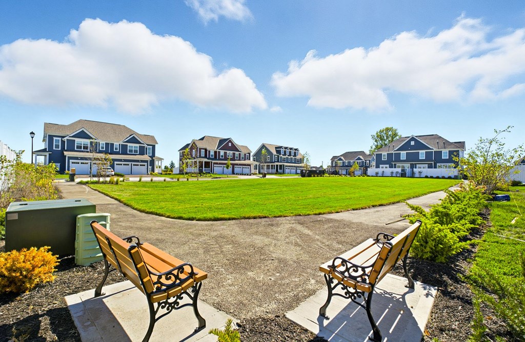 Two benches are placed in a park with a grassy area and houses in the background.