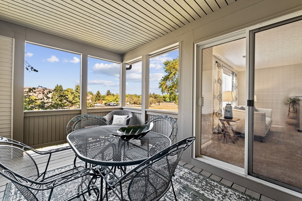 A glass table with chairs is on a patio with a view of trees and a house.