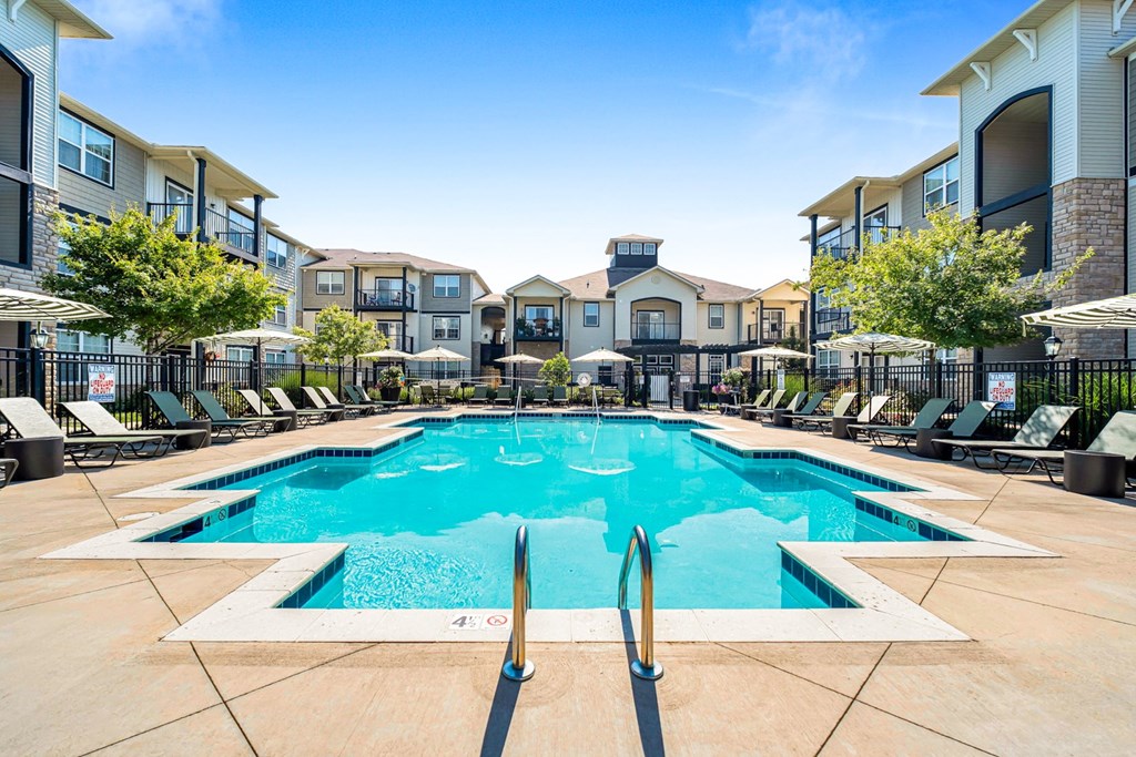 A large swimming pool surrounded by lounge chairs and umbrellas in front of apartment buildings.