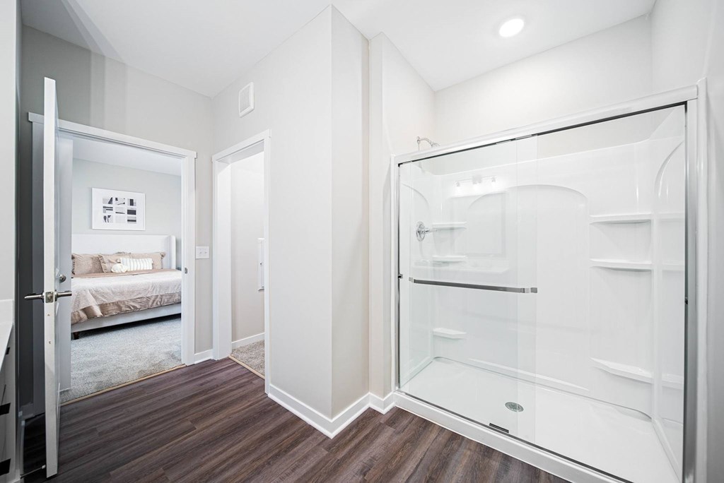 A white bathroom with a walk-in shower and a bedroom visible through the door.
