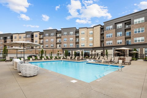 A large swimming pool is surrounded by lounge chairs and umbrellas in front of a large apartment building.