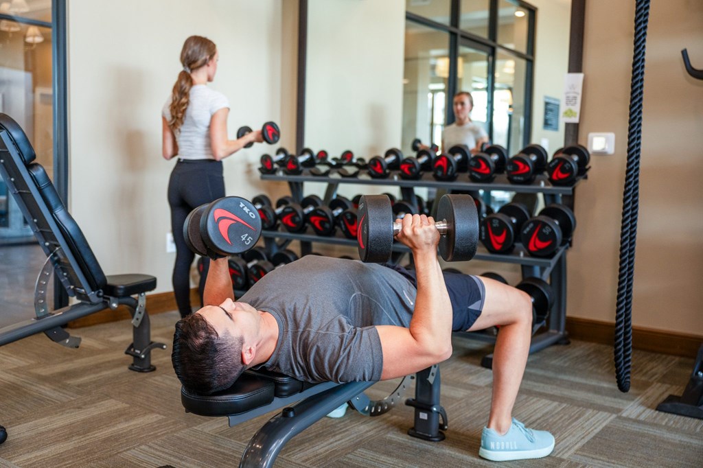 A man is working out in a gym with a woman in the background.