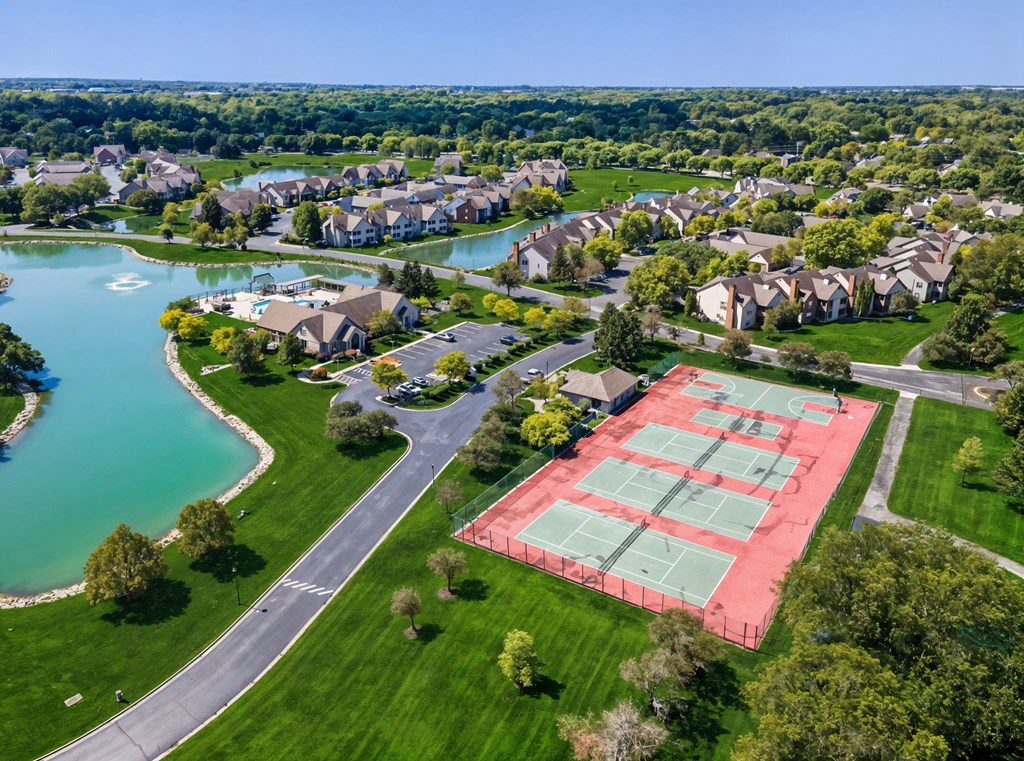 A tennis court is located in a grassy area near a lake and houses.