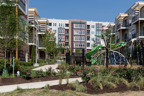 A playground with a green slide and a white swing set is surrounded by a garden with red flowers and green shrubs.