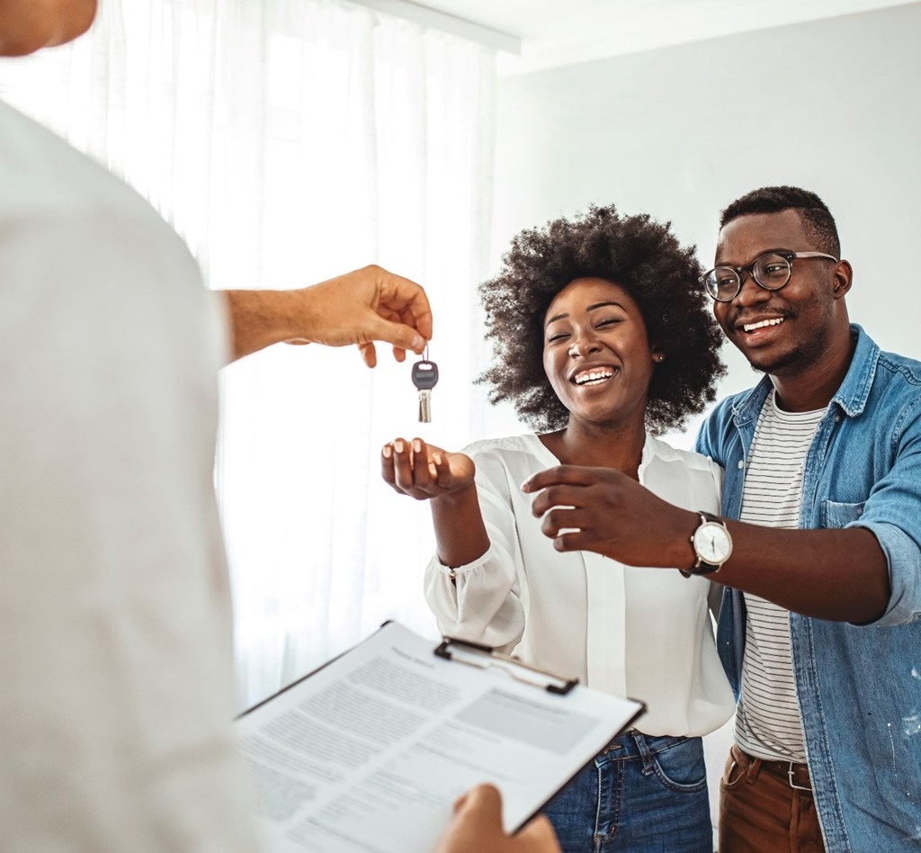 A person is handing over a key to a couple who are smiling and looking at a document.