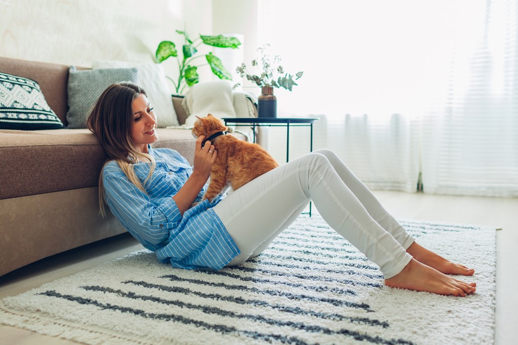 A woman in a blue shirt is sitting on a rug with her orange cat.