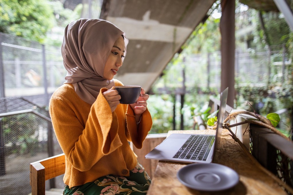 A woman in a yellow top and floral skirt is sitting at a table with a laptop and a cup in front of her.