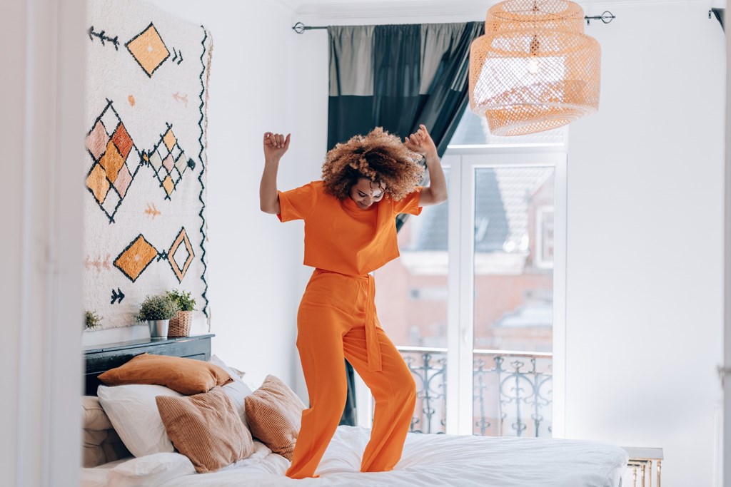 A person in an orange outfit is dancing in a room with a window and a bed.