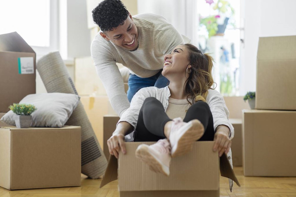 A man and woman are playing in a cardboard box in a room with other boxes and a couch.