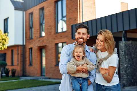 A family of three stands in front of a modern house.
