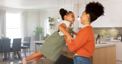 A woman in an orange top is holding a child in a green dress in a kitchen.