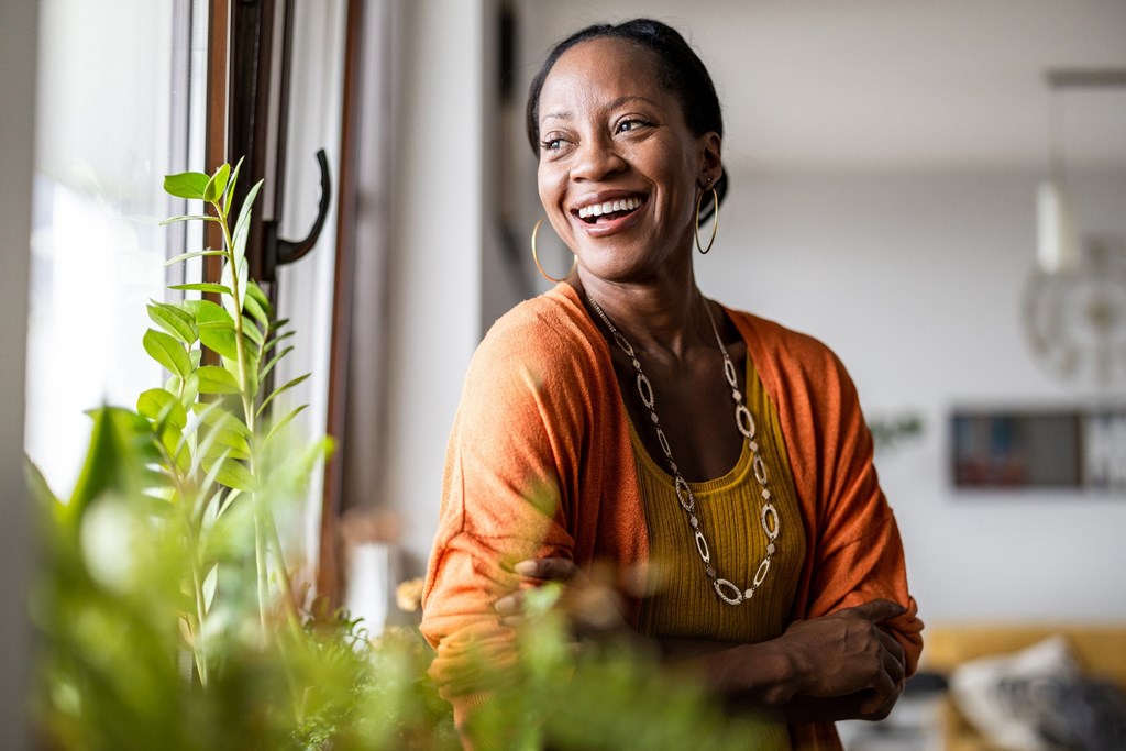 A woman in an orange top is standing in front of a window with a plant beside her.