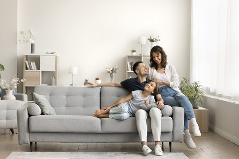 A woman and a child are sitting on a grey couch with a man lying on the floor.