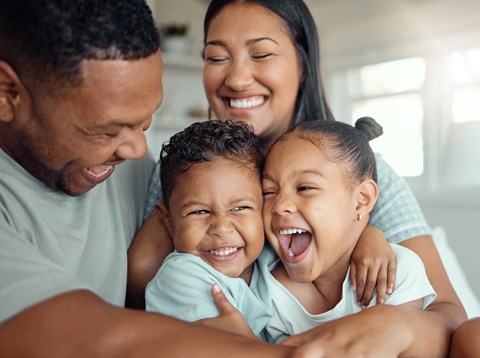 A family of four is hugging and smiling together in a bright room.