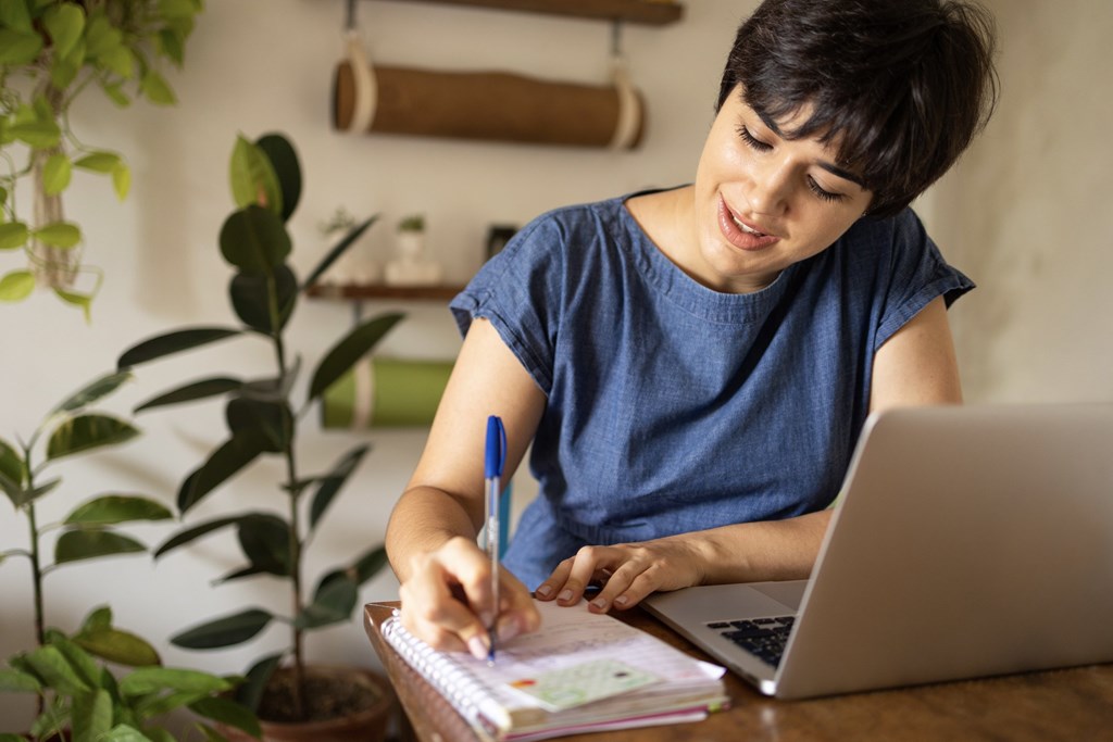A person in a blue shirt is writing in a notebook with a laptop open beside them.