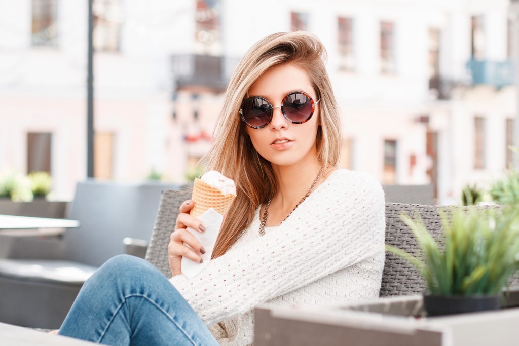 A woman in a white sweater and blue jeans is sitting on a chair holding an ice cream cone.