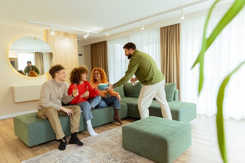 A man serving food to two people sitting on a green couch in a living room.