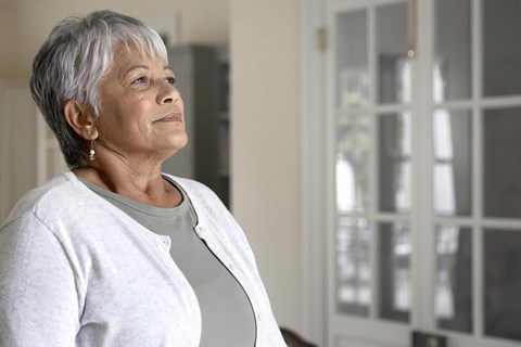 A woman with short grey hair wearing a white cardigan and a grey top is standing in front of a white door.