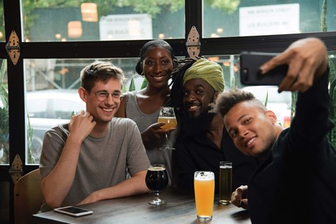 Four people are sitting at a table with drinks and taking a selfie.