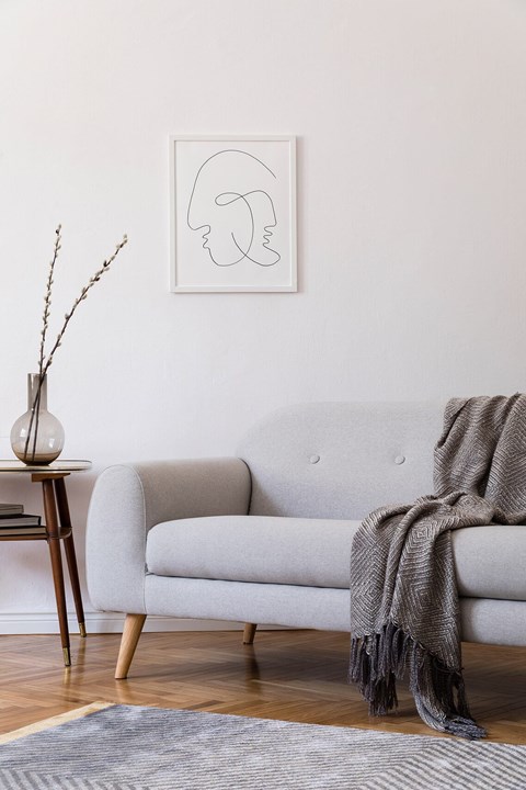 A grey couch with a blanket draped over it sits in a room with a vase on a small table.