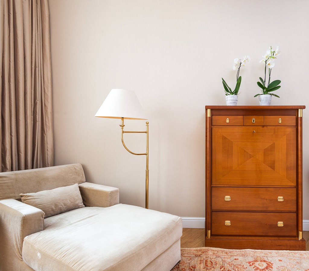 A white sofa sits in front of a wooden chest of drawers with a lamp and two potted plants beside it.