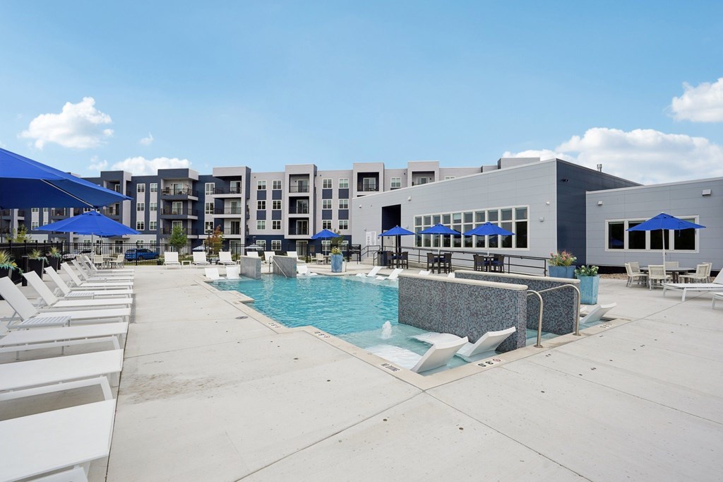 A swimming pool surrounded by lounge chairs and umbrellas in front of apartment buildings.