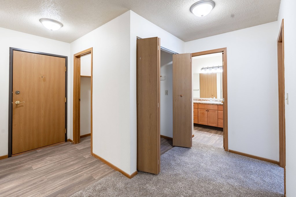 A room with a carpeted floor and white walls with a brown door and a wooden cabinet. Fargo, ND Country Edge Apartments