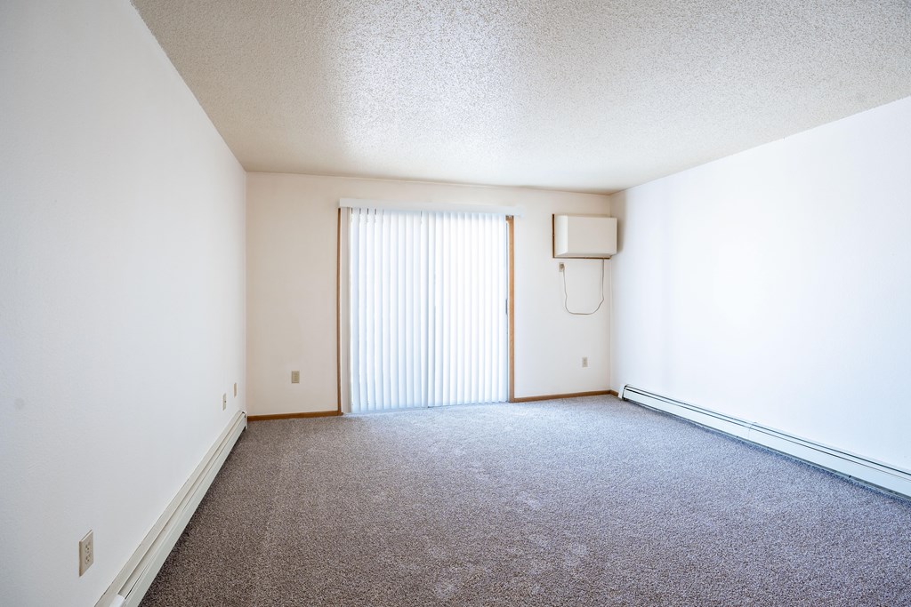 A room with a carpeted floor and a window with blinds. Fargo, ND Country Edge Apartments