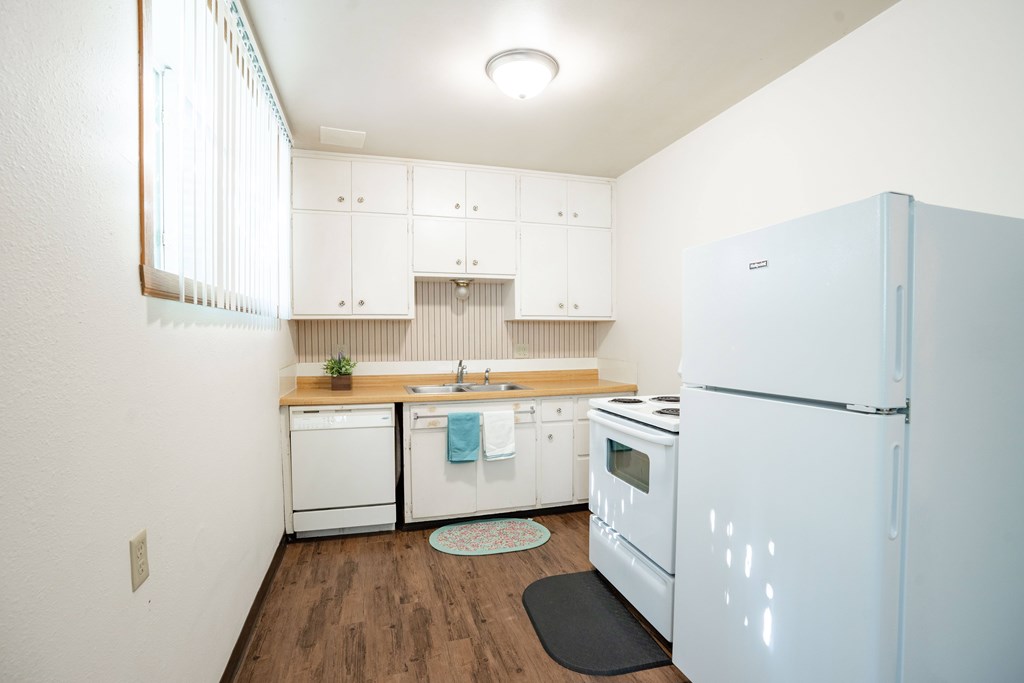 A white refrigerator is in the corner of a kitchen with white cabinets and wooden floors. Fargo, ND L&R Apartments