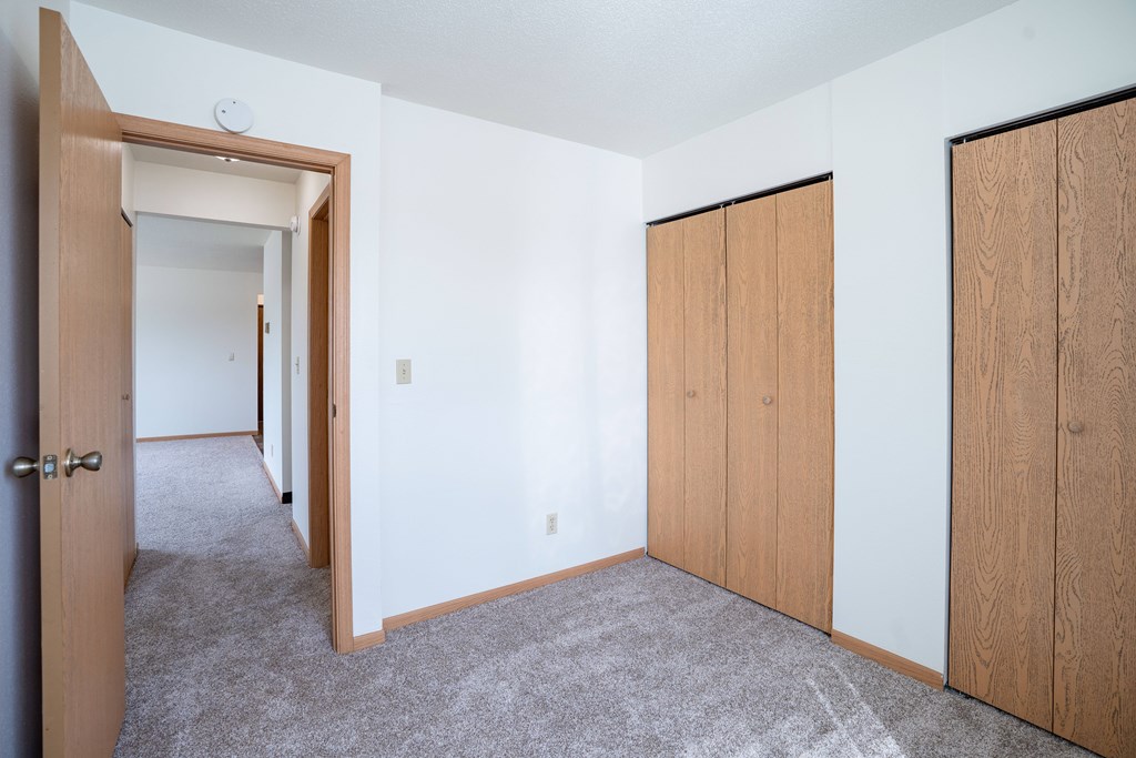 A room with a carpeted floor and a white wall with a door and a window. West Fargo, ND Saddlebrook Apartments