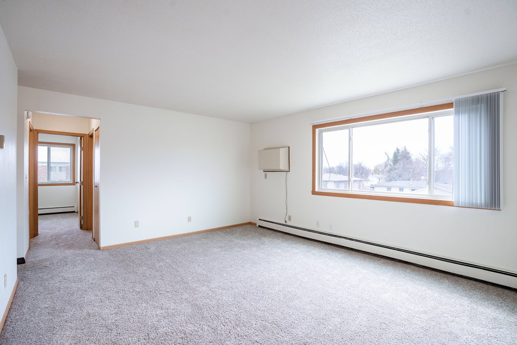 A large, empty room with a carpeted floor and a window with curtains. West Fargo, ND Saddlebrook Apartments