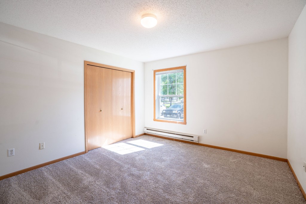 A bedroom with a carpeted floor, a door, a window. Fargo, ND Stonebridge Apartments