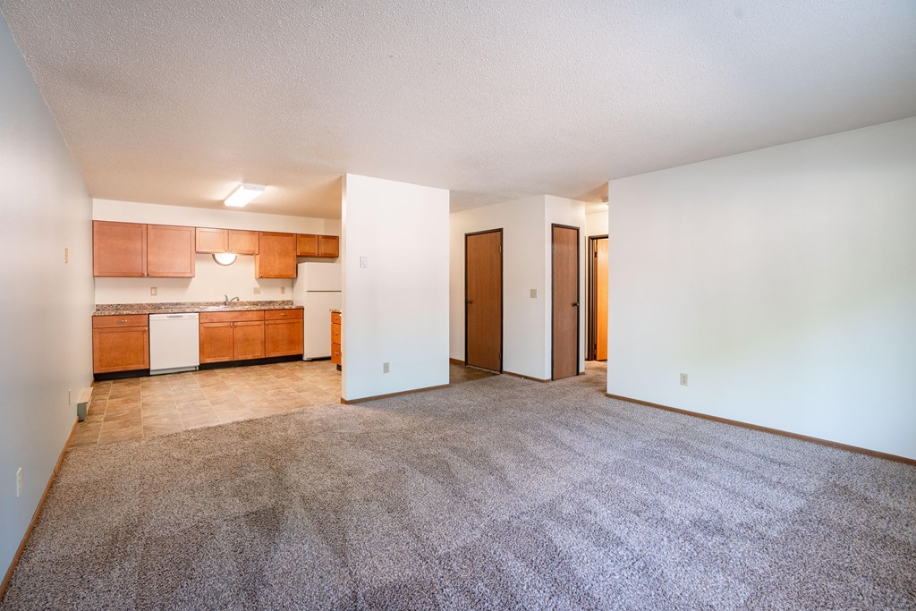 A room with a carpeted floor and a kitchen area in the background. Fargo, ND West Oak Apartments
