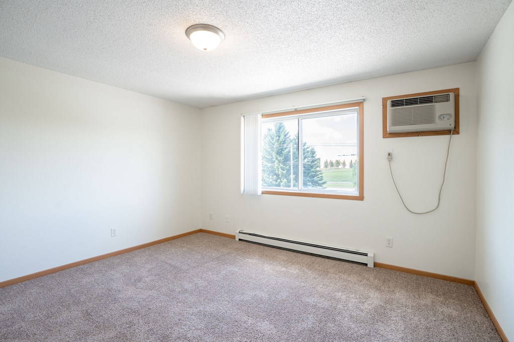 A room with a window and a wall-mounted air conditioner unit. Bismarck, ND Sierra Ridge Apartments