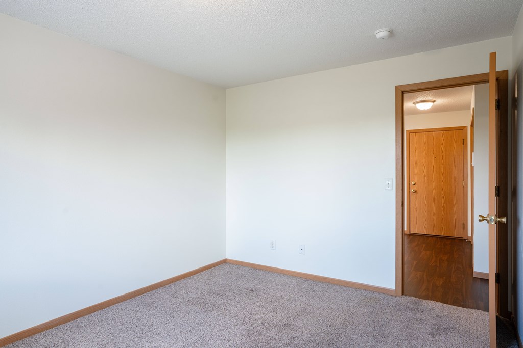 A room with a carpeted floor and a wooden door. Bismarck, ND Sierra Ridge Apartments