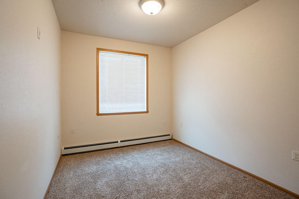 A room with a carpeted floor and a window with blinds. Fargo, ND North Sky II Apartments