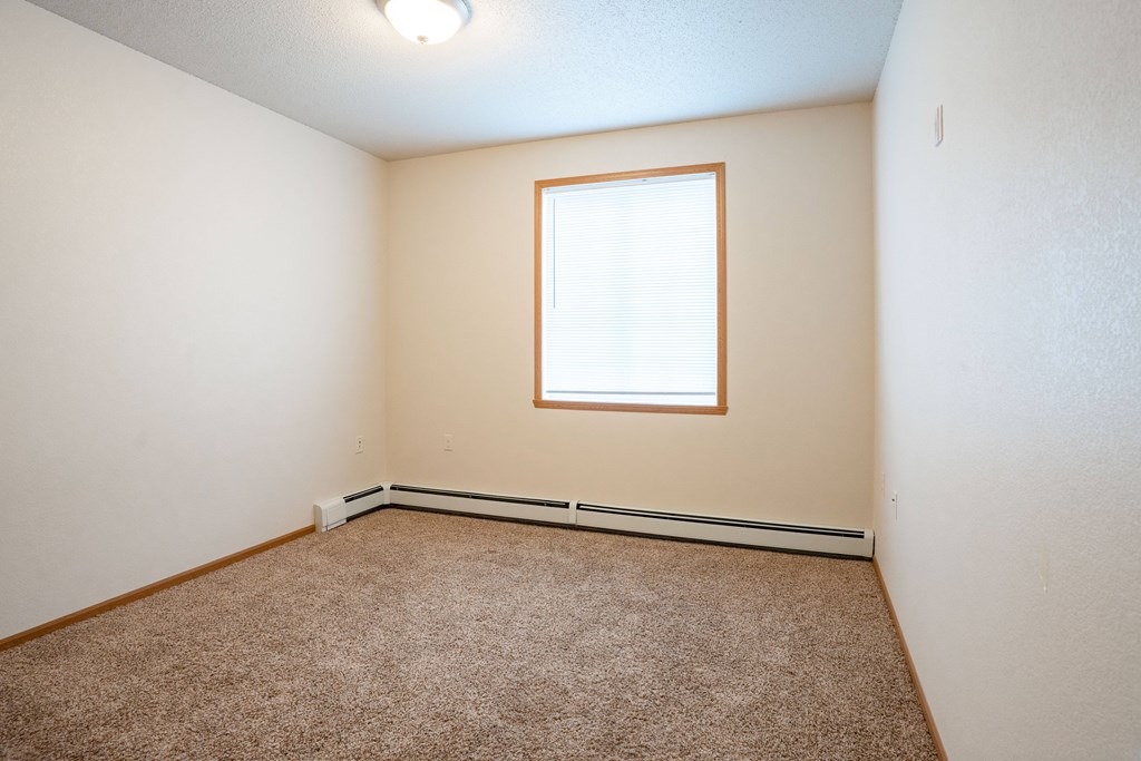 A room with a carpeted floor and a window.. Fargo, ND North Sky II Apartments