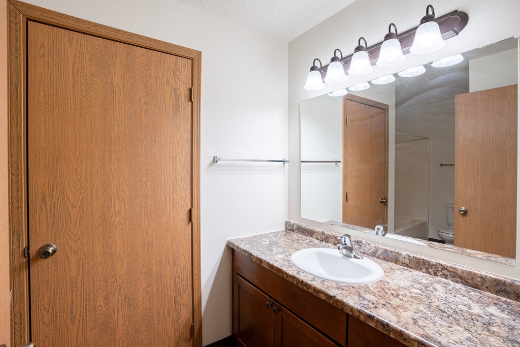 A bathroom with a sink, mirror, and wooden door. Fargo, ND Park Place Apartments