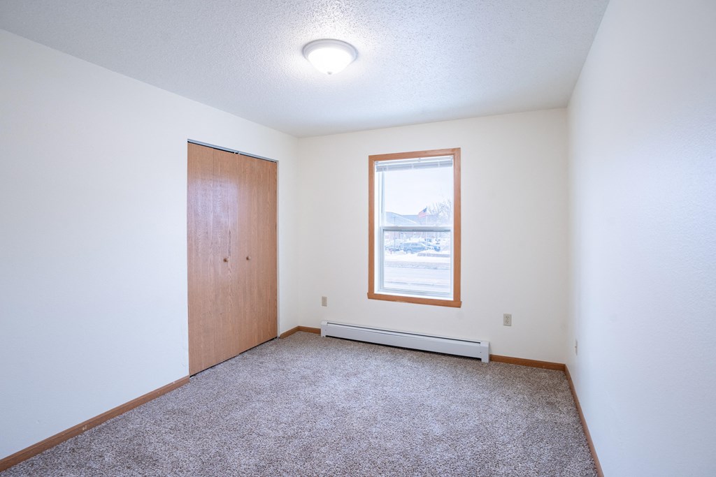 A room with a carpeted floor, a door, a window, and a small radiator. Fargo, ND Park Place Apartments