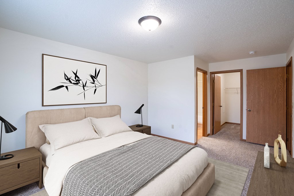 A bedroom with a bed, nightstand, lamp, and a framed picture on the wall. Fargo, ND Park Place Apartments