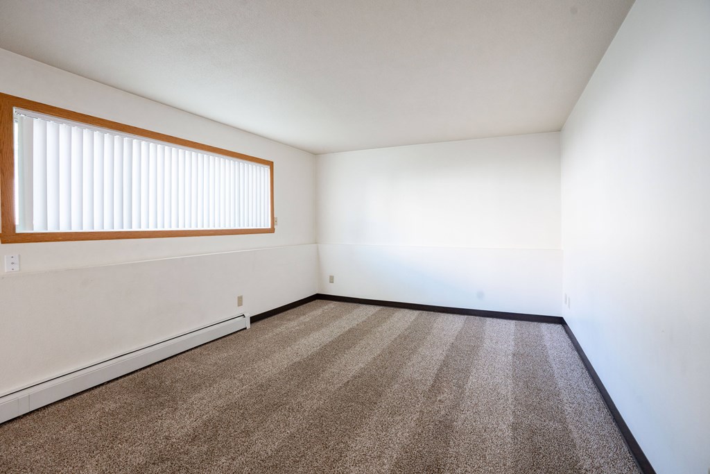 A room with a carpeted floor and a window with blinds. Fargo, ND Islander Apartments