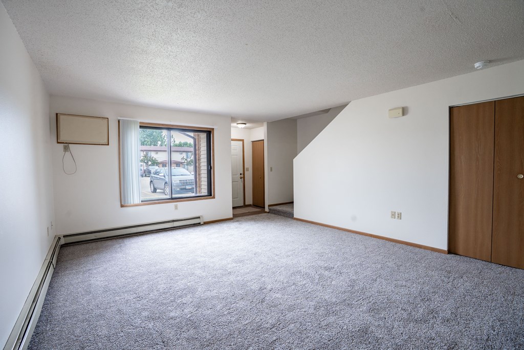 A room with a grey carpet and a window with a view of trees and buildings outside. Fargo, ND Hazelwood Townhomes