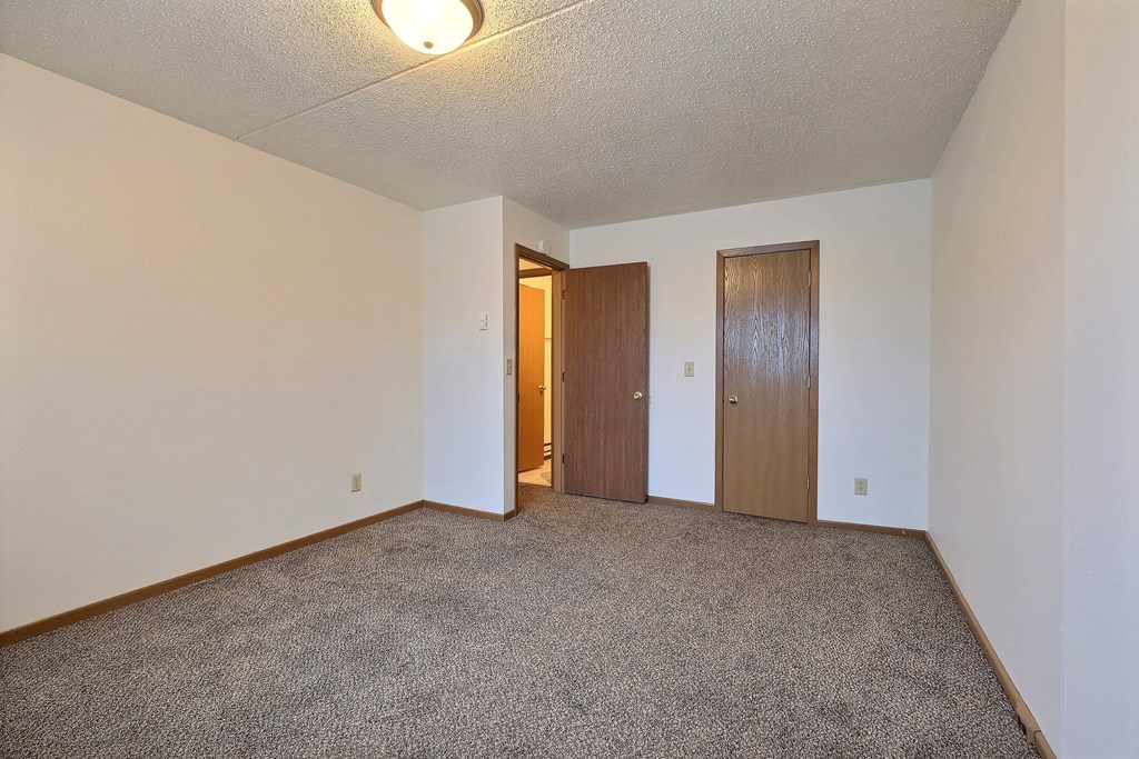 a spacious bedroom with carpet and a door to a hallway. Fargo, ND Alden Pines Apartments